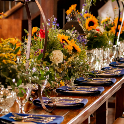 Table de réception élégamment dressée avec des compositions florales champêtres à base de tournesols, roses et feuillages, accompagnées de serviettes bleues et couverts raffinés, dans une ambiance chaleureuse et rustique. decoration-table-reception-fleurs-champetres-tournesols
