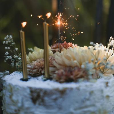 Gâteau de fête décoré de fleurs fraîches et de gypsophile, orné de bougies dorées allumées et d’une étincelle scintillante, créant une ambiance élégante et festive idéale pour un anniversaire ou un mariage. gateau-fleurs-bougies-etincelle-decoration-festive-anniversaire