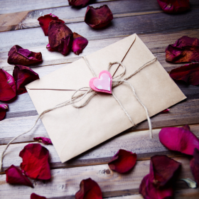 Lettre d’amour enveloppée dans du papier kraft, nouée avec une ficelle et décorée d’un cœur rose, posée sur un lit de pétales de rose rouge. lettre-amour-romantique-petales-roses