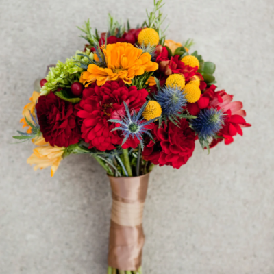 Bouquet de fleurs colorées aux tons chauds composé de dahlias rouges, de craspedias jaunes, de chardons bleus et de fleurs orange, lié par un ruban beige, parfait pour un mariage ou une décoration automnale. bouquet-fleurs-couleurs-chaudes-automne-mariage