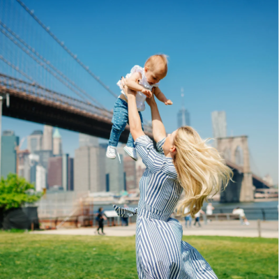 Mère jouant avec son bébé dans un parc ensoleillé, avec le pont de Brooklyn et la skyline de New York en arrière-plan. mere-bebe-new-york-pont-brooklyn-plein-air