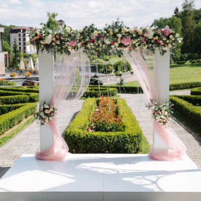 Arche florale de mariage décorée de pivoines roses et blanches, de feuillages et de voilages roses, installée dans un jardin élégant avec allées de buis taillés et fontaine en arrière-plan. arche-mariage-pivoines-decoration-jardin