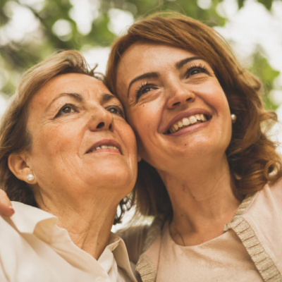 Portrait d’une mère et sa fille souriantes, partageant un moment complice en extérieur sous les arbres. mere-fille-moment-complice-plein-air
