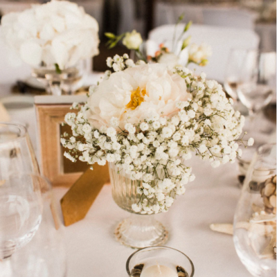 Centre de table de mariage élégant avec une pivoine blanche entourée de gypsophiles, présenté dans un vase en verre sur une table décorée avec bougies, verres et éléments raffinés. centre-table-mariage-pivoine-gypsophile-decoration-elegante.jpeg