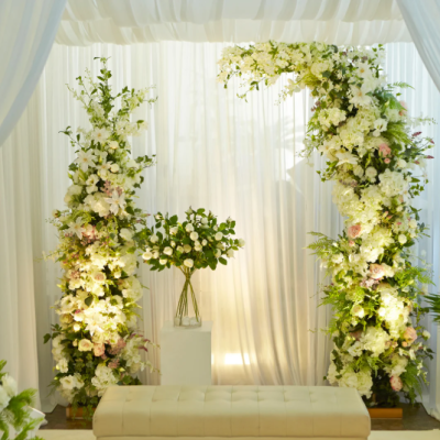 Décoration florale de cérémonie de mariage en intérieur, avec une arche élégante ornée de fleurs blanches et verdure, accompagnée d’un rideau blanc drapé et d’un banc crème pour une ambiance raffinée et romantique. arche-mariage-interieur-fleurs-blanches-decoration-elegante.png